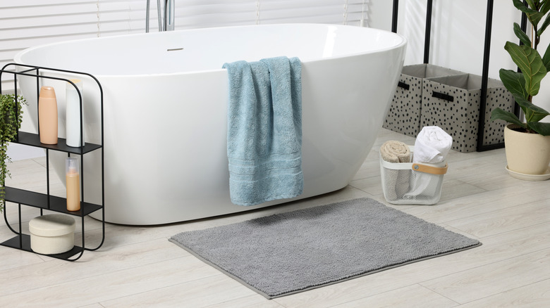 A stand-alone bathtub with a light blue towel draped over the edge and a gray bathmat on the floor
