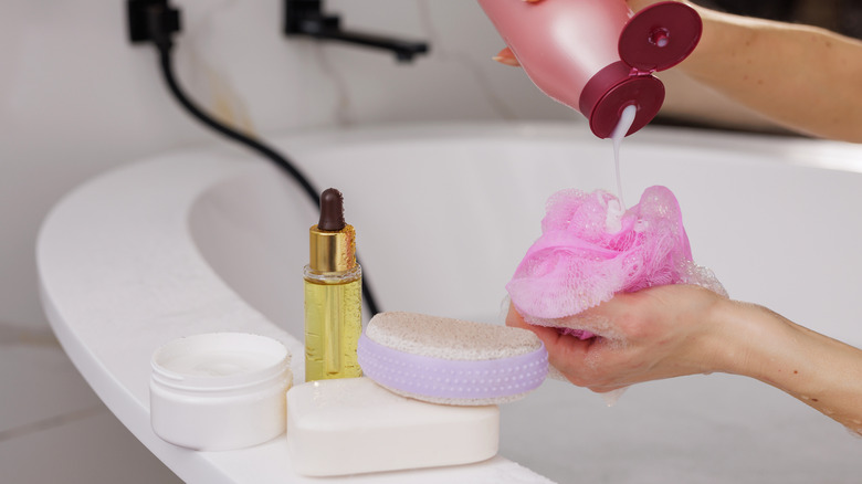 Bath products on a tub's edge with a woman pouring shower gel on a loofah