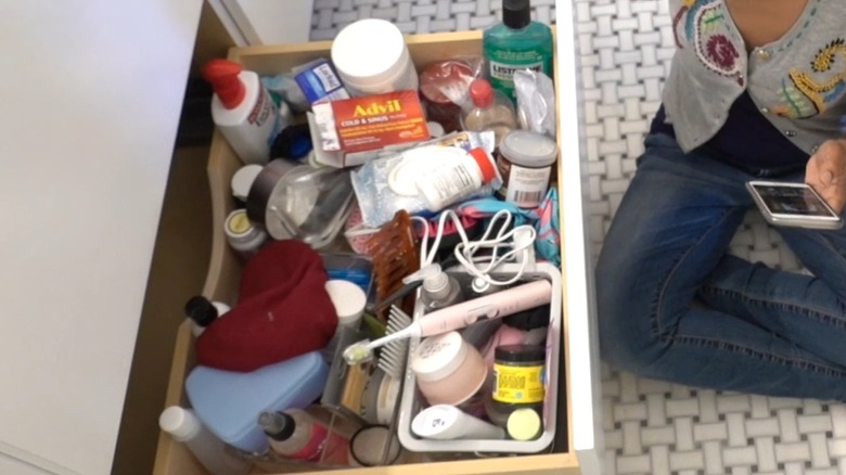A woman sitting beside an open messy drawer in a bathroom