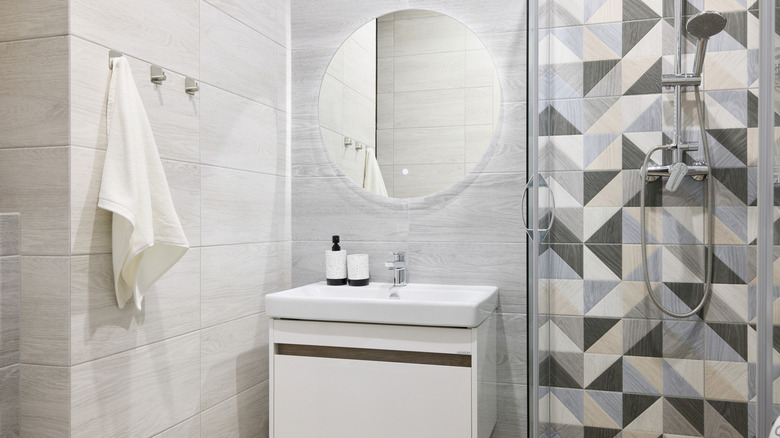 A shower with geometric tiling in black, grey and cream next to a sink and vanity unit with a large circular mirror on the wall above it.