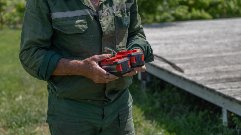 A person holds two rechargeable batteries in their hands while standing in their backyard near a wooden deck.