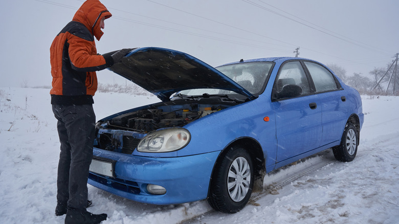 person looking under hood of broken down car in snow