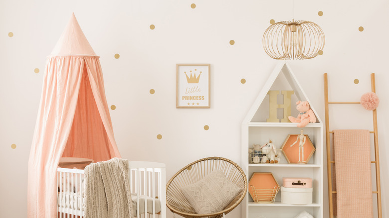 A white crib with a salmon fabric canopy sits in front of a cream colored wall with small gold polka dots