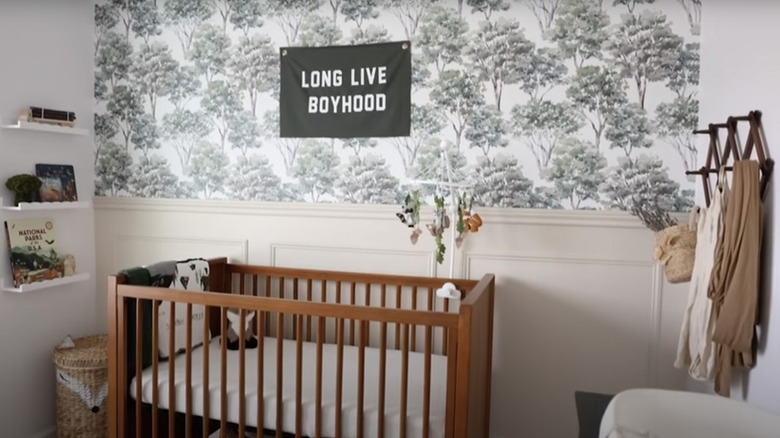 A sign that reads "Long Live Boyhood" hangs over a wooden crib on a wall covered in toile print wallpaper