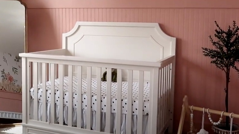 A white crib sits in front of an accent wall covered in pink beadboard