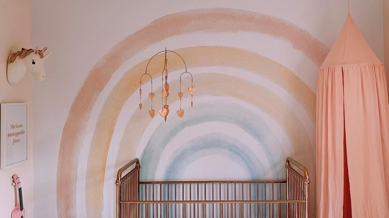A soft watercolor rainbow mural is behind a metallic crib and baby mobile