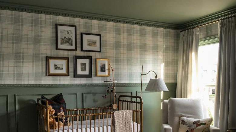 A brushed gold crib sits in front of a wall covered with plaid wallpaper and green decorative trim