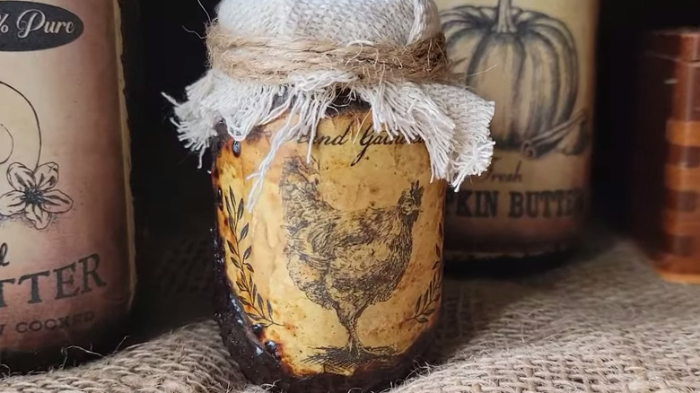 A mason jar with a dark and chunky outside and a label that appears old with the top covered in burlap and twine