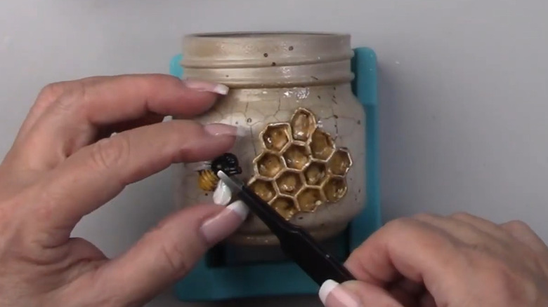 A person using tweezers to attach a bee to a jar with fine line cracks and a gold beehive