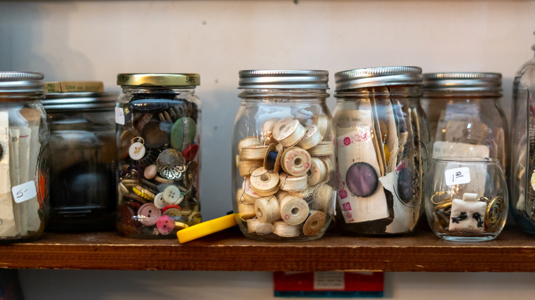 A series of mason jars full of random trinkets and items including buttons and thread on a shelf
