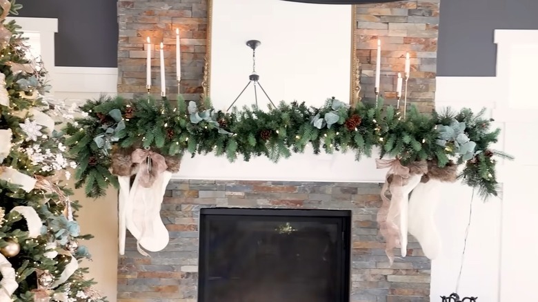 Thick evergreen garland with lights is accented with white candles and stockings on a mantel.
