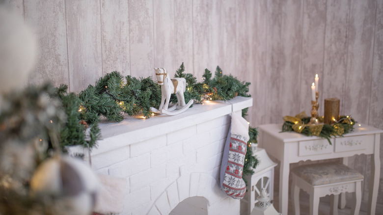 A simple lighted evergreen garland, rocking horse, and stocking decorate a white wooden fireplace.