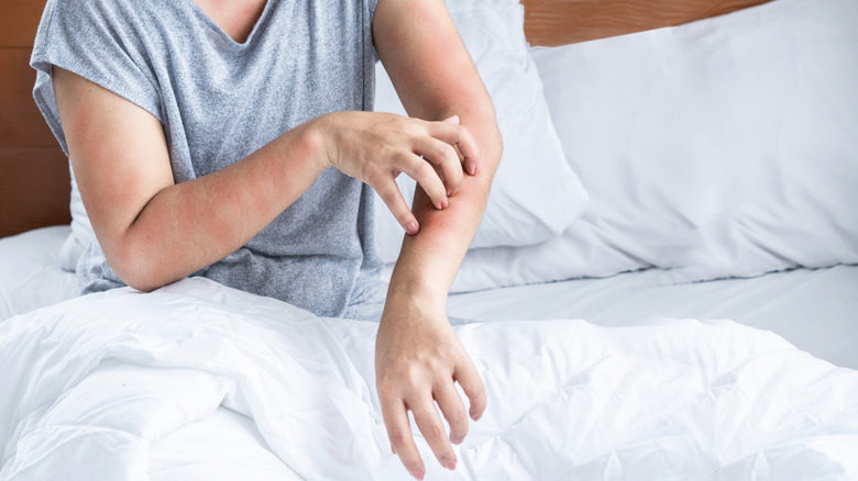 A person sits in bed scratching their arm due to an infestation of bugs.