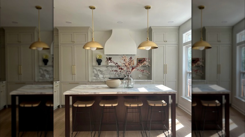 Image of an airy kitchen with gold pendant lights, hearth style range hood, marble countertops, and kitchen island