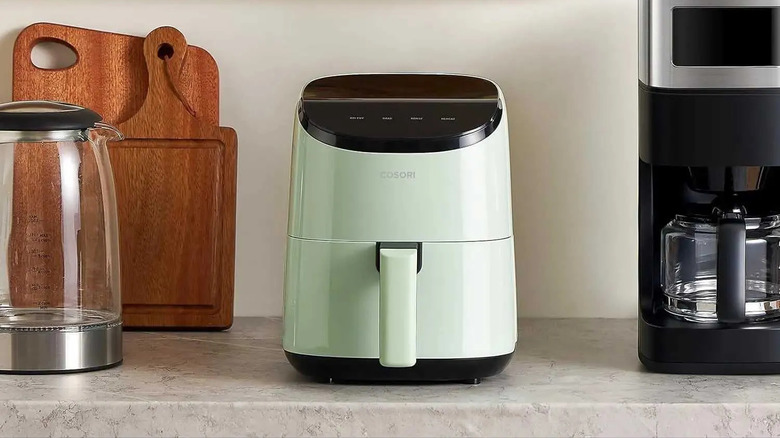 A mint green Cosori Lite Series Air Fryer sits on a gray countertop next to a coffeepot and wooden cutting boards