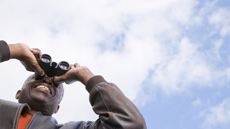 A smiling Black man looks through binoculars against a partly cloudy sky.