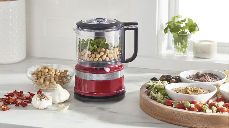 A red KitchenAid mini food processor on a table with garlic, tomatoes and olives.