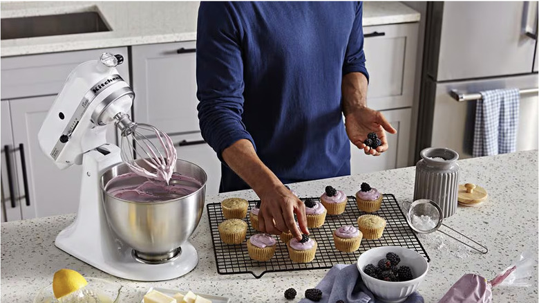 A black KitchenAid stand mixer sits on a countertop between peeled apples and an apple pie.