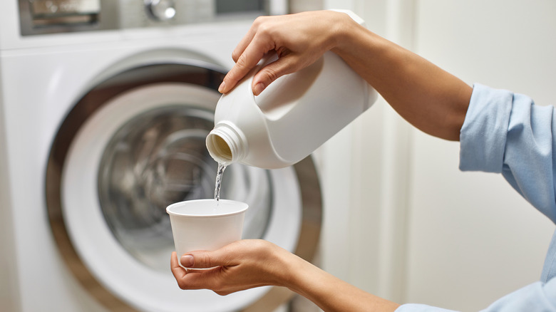 A person measuring out bleach in a cup in front of a washing machine