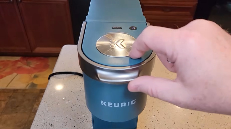 Person reaching to open the top of a blue Keurig machine