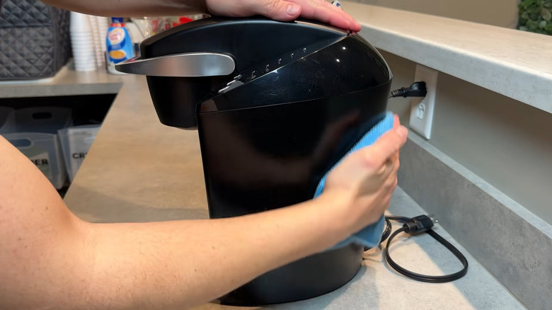 Person wiping the exterior of a black Keurig machine with a blue cloth