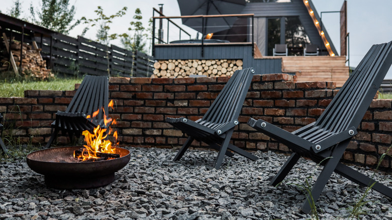 Burning firewood in a metal firepit surrounded by gravel and black wooden chairs, with a modern A-frame cabin and string lights in the background at twilight.