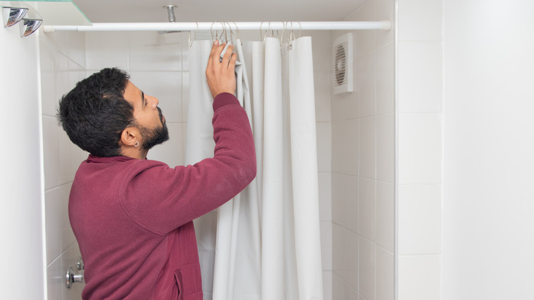 A man with dark hair and a short beard, wearing a long-sleeved maroon shirt, stands in a white bathroom adjusting a white shower curtain.