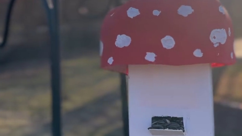 A mushroom-shaped upcycled milk jug bird feeder hanging on a pole in a yard