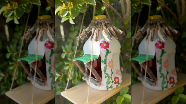 A milk jug bird feeder with sticks holding a flap open for an entry way with a stick perch and stick roof on a hanging shelf