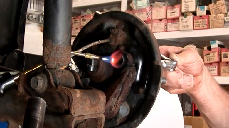 Person using blowtorch on rusted car bolt while partner uses socket wrench