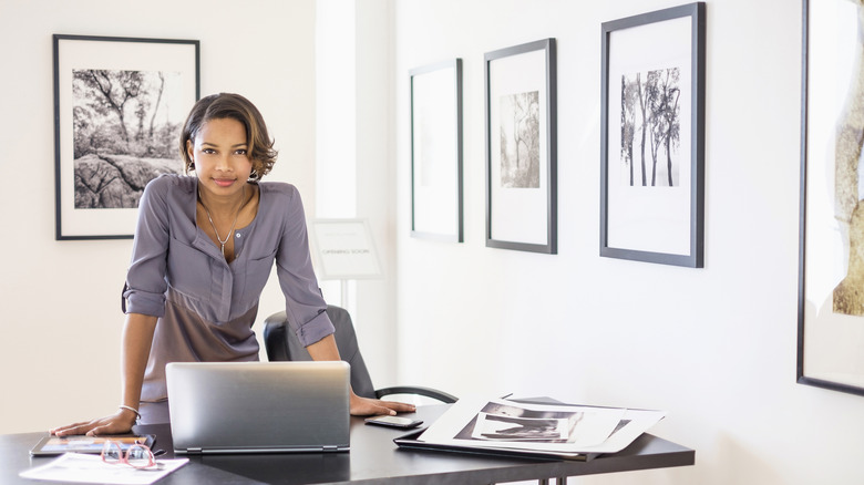A woman stands in front of a desk with a laptop and other items as different framed portraits are hung on the walls behind her.