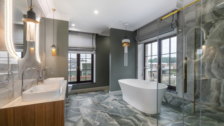 An open bathroom with gray marble, a standalone bathtub, a shower, two sinks, and a semi-separate toilet