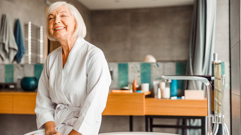 A smiling older woman in a bathrobe sits on the edge of a soaking tub