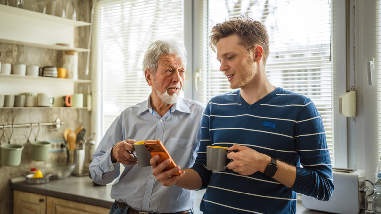 An older man speaking with a young man holding a phone while drinking coffee in the kitchen