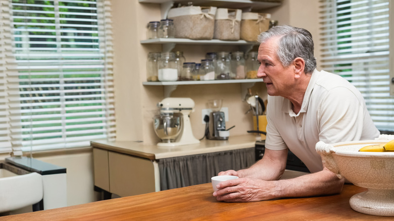 A senior man drinks coffee while leaning on wood counter, with open shelving in background