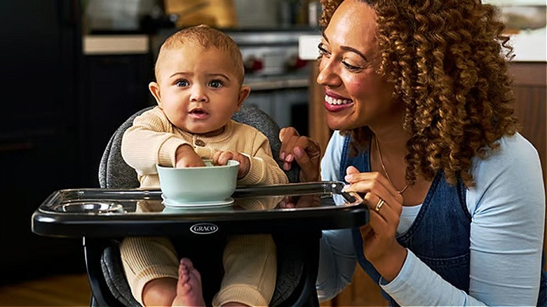 A toddler sitting in a high chair while a happy mother looks on