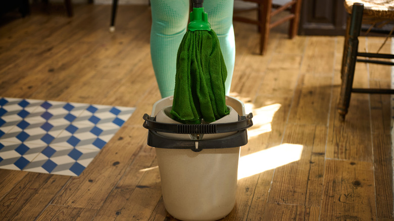 A person stands behind a bucket and mop on a wooden floor