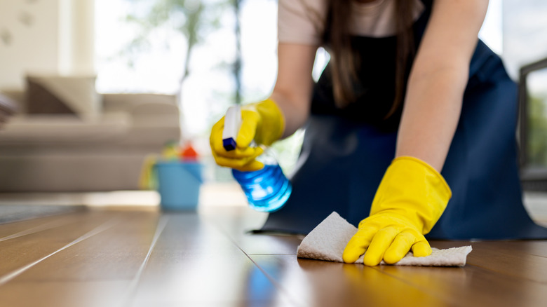 A person wearing yellow gloves cleans a wooden floor with spray