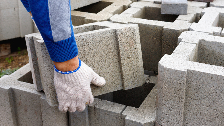 A person with gloved hands lifts a gray concrete block from a stack of other blocks.