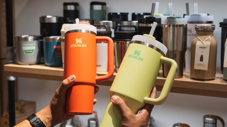 A person holds two Stanley cups in front of a shelving unit packed with tumblers