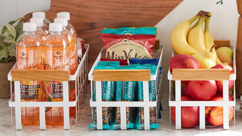 A set of three Wire Pantry Bins by Member's Mark full of fruits, snacks, and drinks, side-by-side on a counter
