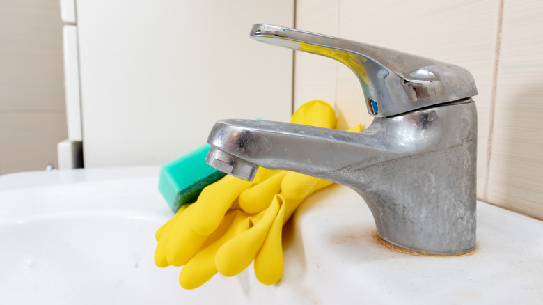green sponge and yellow gloves sitting on sink near dirty faucet with limescale and calcified water on tap