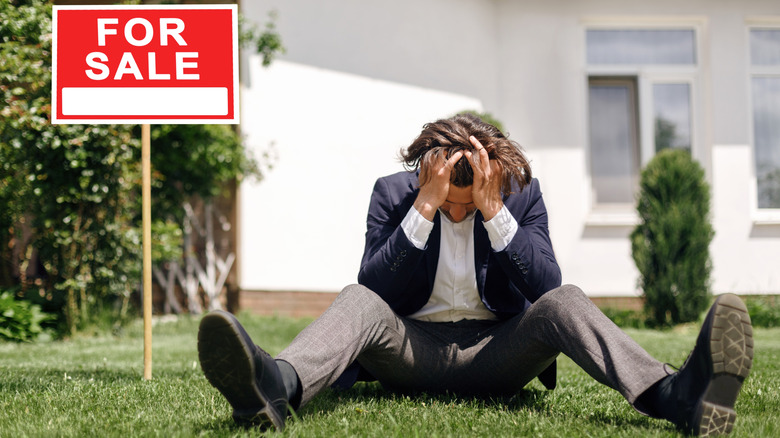 upset man sitting in the grass in front of a house with a for sale sign