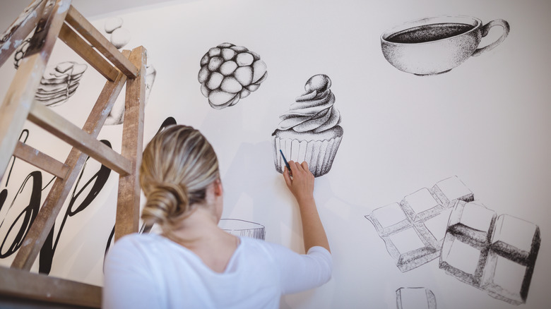 Woman painting black and white food murals on a kitchen wall with a ladder