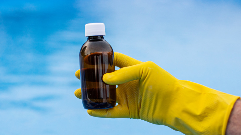 A person holding a dark glass bottle with yellow gloves in front of a pool.