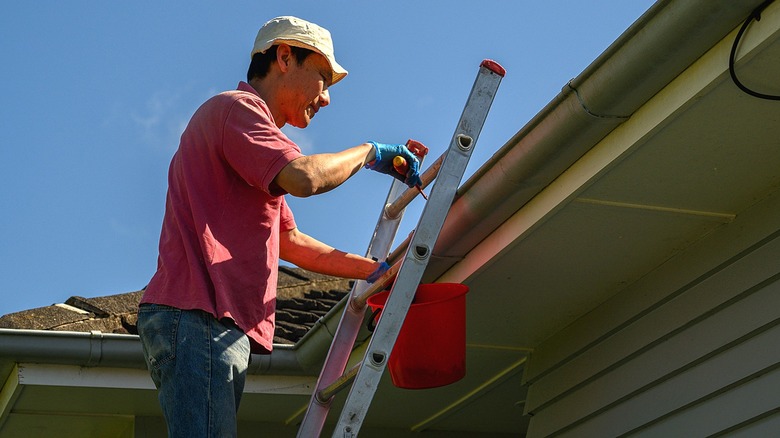 A person standing on a ladder and holding a bucket to clean gutters