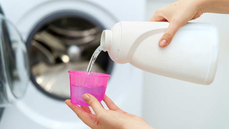 Laundry detergent pouring from a white bottle into a pink cap with washing machine behind.