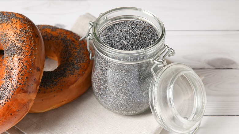 A jar of poppy seeds sitting open next to two poppy seed bagels.