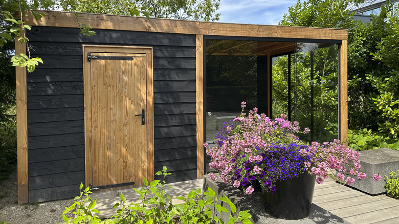 A large planter full of colorful flowers sits in front of a modern black shed