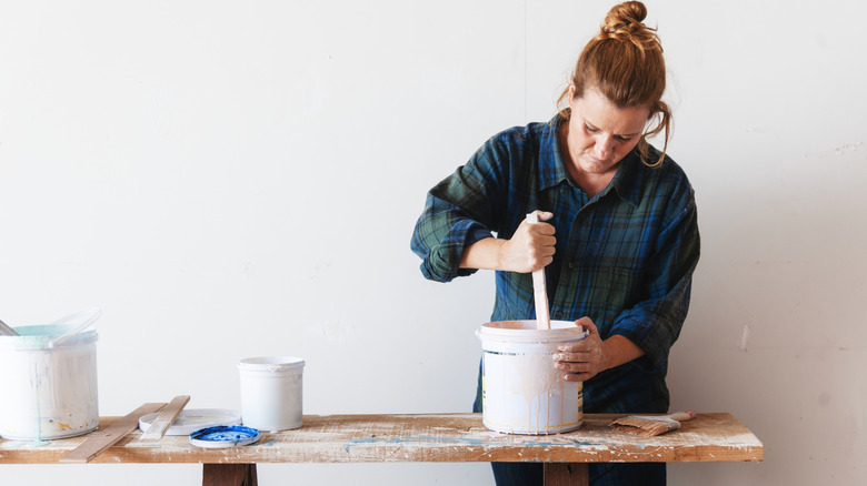 A woman mixing paint in a bucket on a wooden table with painting supplies